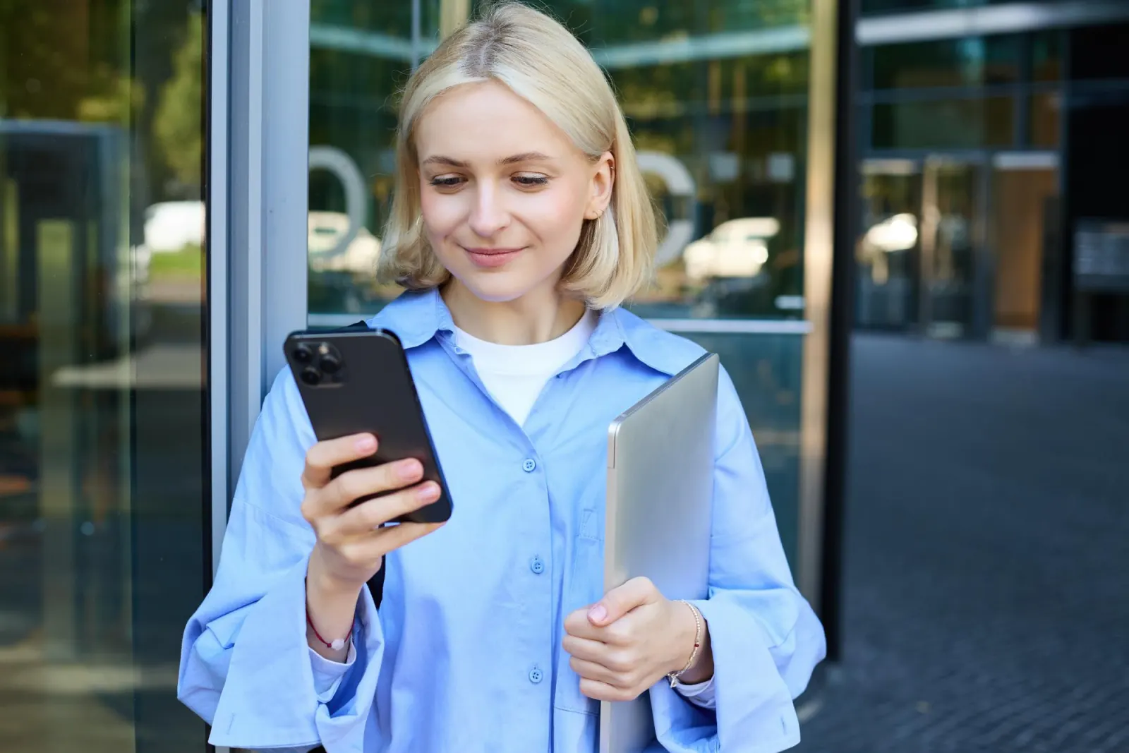 Woman holding a laptop, smiling as she checks messages on her phone outside an office